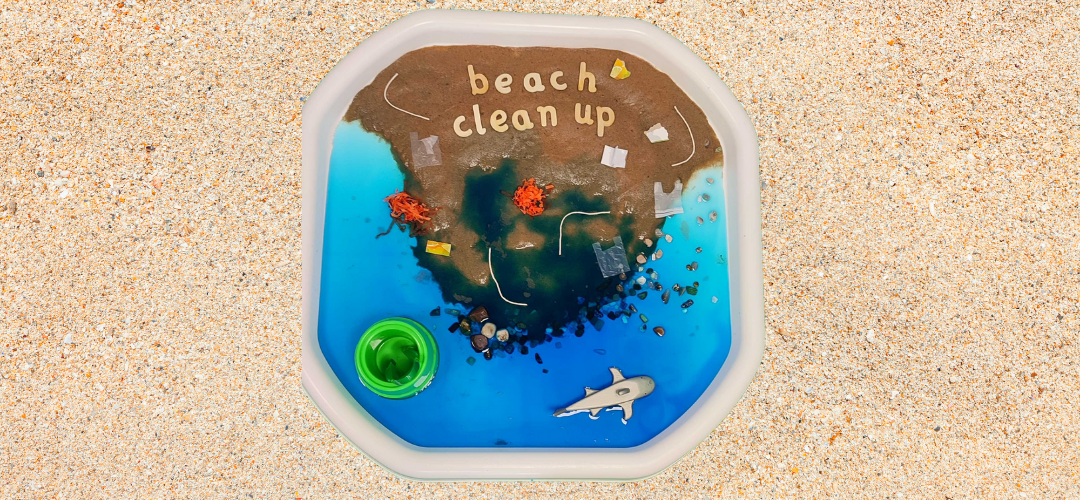 overhead shot of a tuff tray with a beach clean up scene. Half of the tray is covered in sand and the other half has water dyed blue. There are small craft pieces designed to look like rubbish on the beach