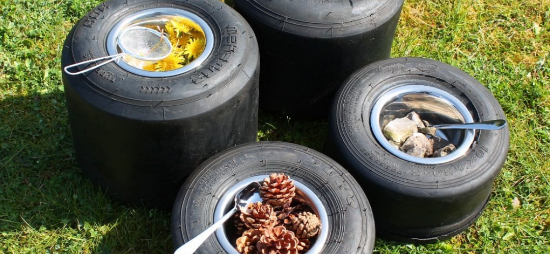 image shows 4 small tyres with metal bowls and utensils placed inside. Each bowl is filled with natural objects, pine cones, pebbles and flowers