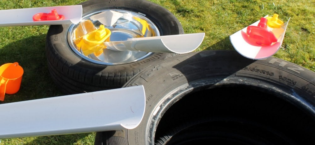large car tyres with white guttering balanced on the side, some guttering leading into other tyres with metal bowls placed inside and toy boats on the guttering