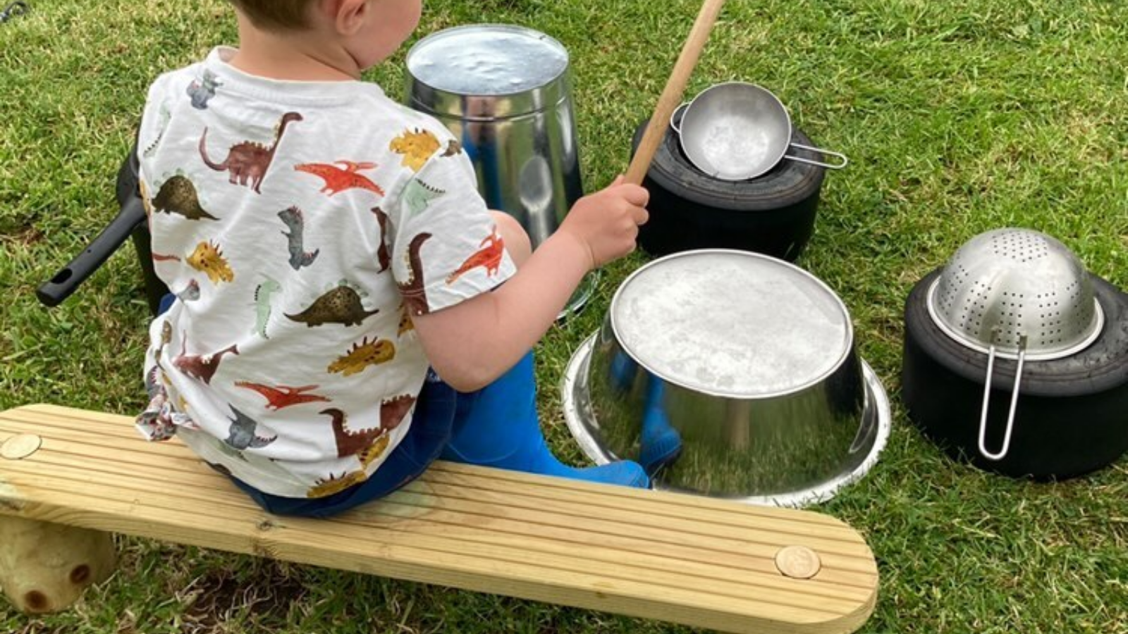 Child sat on a plank with metal pots and pans in front of him, some propped up on small tyres - creating a drum kit set up