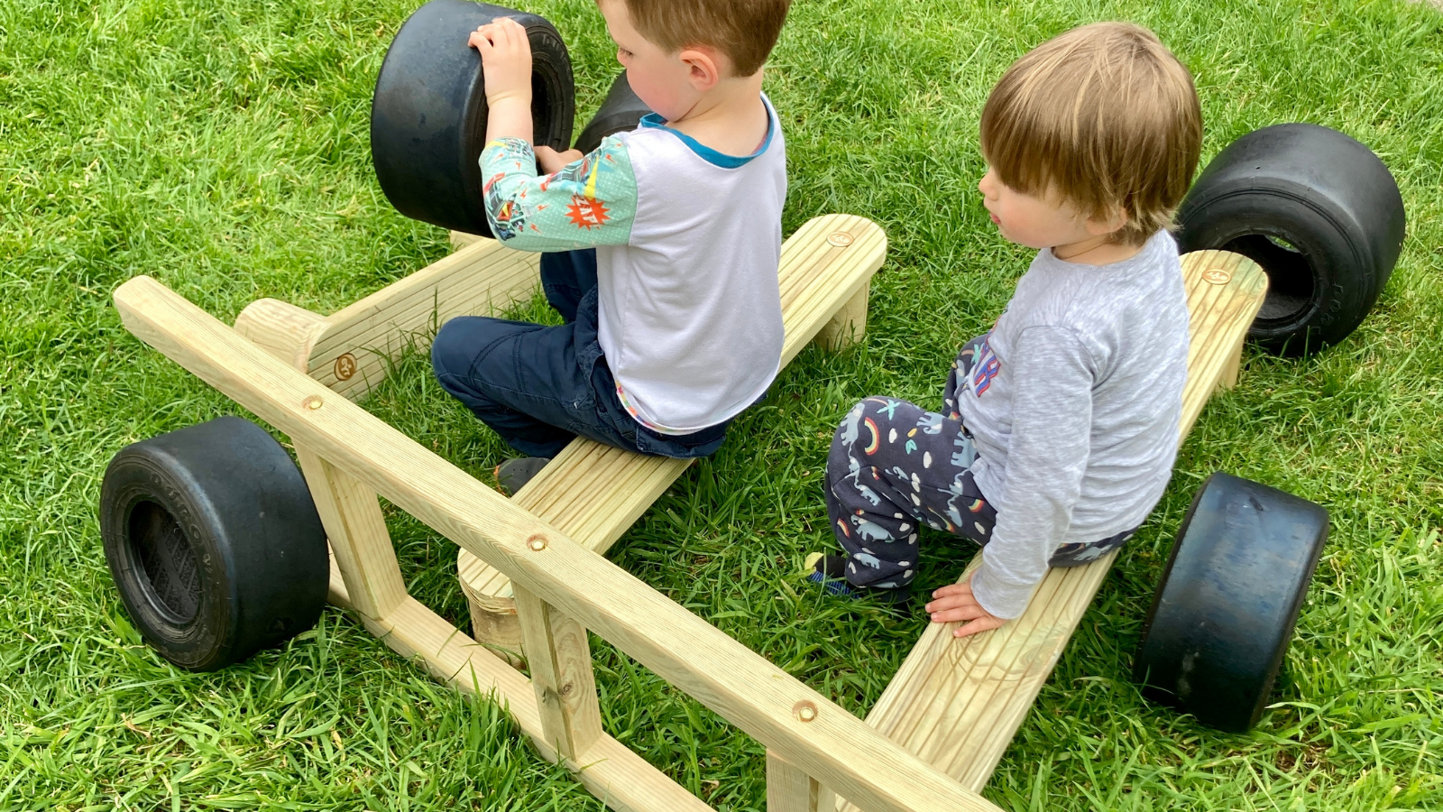 children sat on wooden planks using small tyres as a steering wheel and other planks arranged around to create a car structure